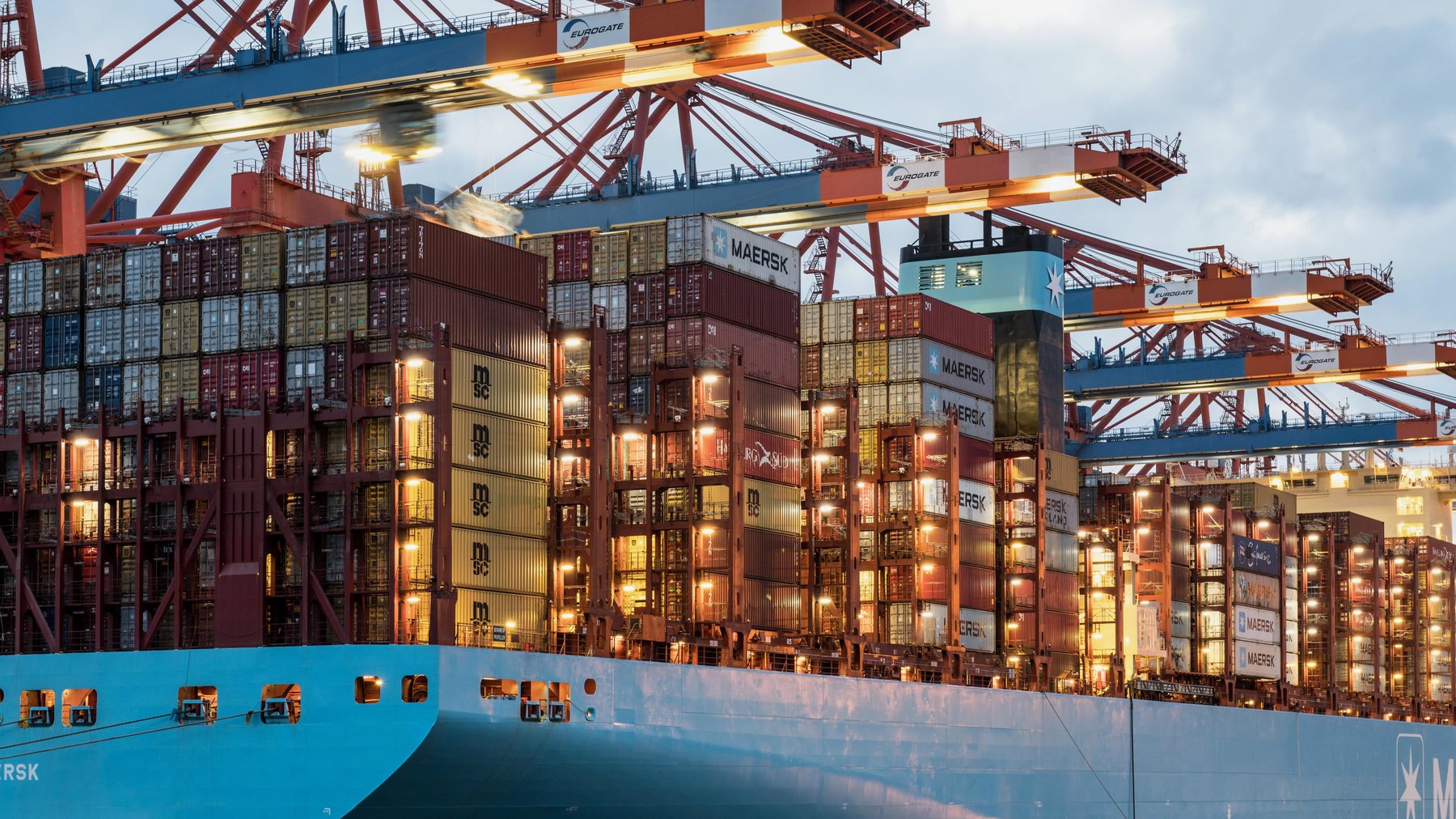 Container ship loading at a busy port at dusk with cranes and stacked containers